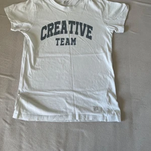 Vit t-shirt med tryck - Säljer en vit t-shirt med texten 'CREATIVE TEAM' i grått. T-shirten har en klassisk rund hals och korta ärmar. Perfekt för en avslappnad stil. Hör av dig vid intresse!