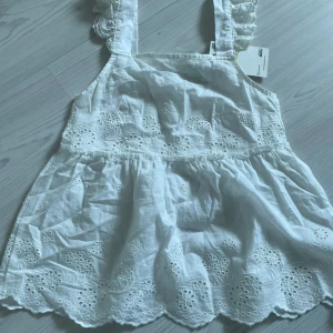 Vitt linne med broderi - Charmigt linne med vackra broderade detaljer och volanger på axelbanden. Perfekt för sommarens varma dagar. 