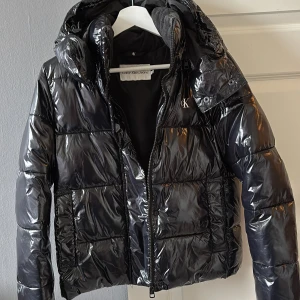 Svart blank jacka från Calvin Klein Jeans - Snygg svart jacka från Calvin Klein Jeans med glansig finish. Jackan har en dragkedja framtill och en huva för extra skydd. Perfekt för kyligare dagar med sin stilrena design och varma vaddering. Finns ett dolt blixtlås om man önskar ta bort luvan.