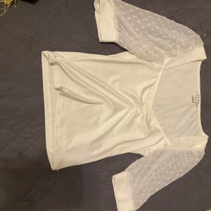 Vit topp med prickiga ärmar - Säljer en stilren vit topp med unika prickiga trekvartsärmar i ett transparent material. Toppen har en v-ringad design och en lätt rynkad detalj framtill. Perfekt för en chic och elegant look.