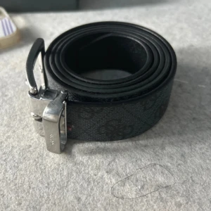 Svart bälte från Guess - Snyggt svart bälte från Guess med ett diskret mönster och en stilren silverfärgad spänne. Tillverkat i Italien, perfekt för att ge en elegant touch till din outfit. Bältet har en klassisk design som passar till många olika stilar.