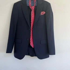 Mörkblå kostym med röd slips från Dressmann - Säljer en stilren mörkblå kostym från Dressmann i slim fit.  Inkluderar även en vit skjorta, slips och näsduk.  Storlek kavaj 42, storlek byxa 29/30, storlek skjorta xs