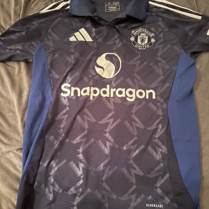 Mörkblå fotbollströja från Adidas - Säljer en mörkblå fotbollströja från Adidas med ett subtilt mönster och vit text. Tröjan har korta ärmar och en krage. Den har ett tryck med 'Garnacho' och nummer 17 på ryggen. Perfekt för fotbollsfans!