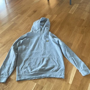 Grå hoodie från Lyle & Scott - Säljer en stilren grå hoodie från Lyle & Scott med deras ikoniska logga på bröstet. Tröjan har en klassisk känguruficka och en bekväm huva. Perfekt för en avslappnad stil. Stl 15-16 år 