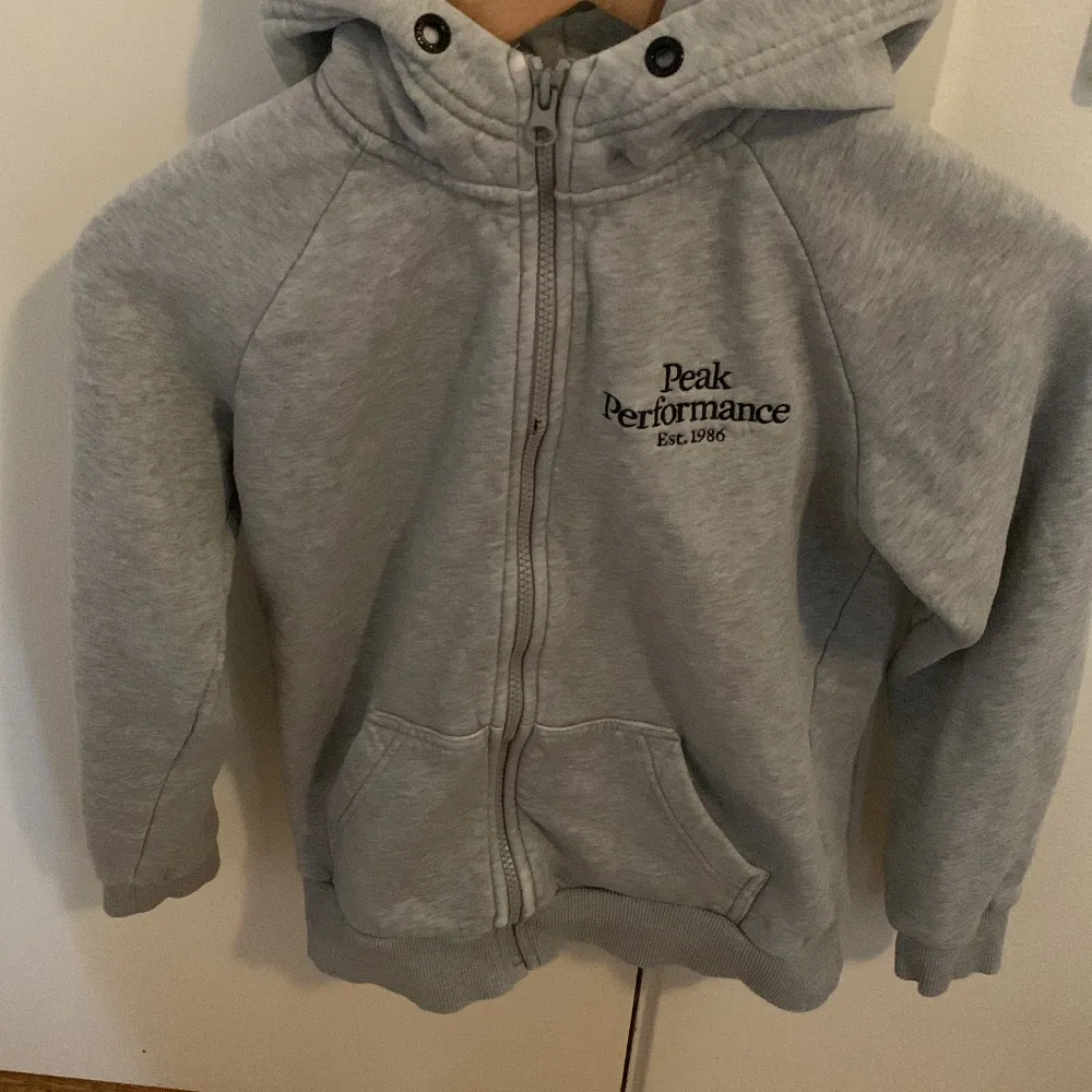 Säljer en grå hoodie från Peak Performance med dragkedja och huva. Tröjan har två fickor framtill och ribbade muddar vid ärmslut och nederkant. Perfekt för en avslappnad stil. Leverans ingår.. Neuletakit & Villapaidat.