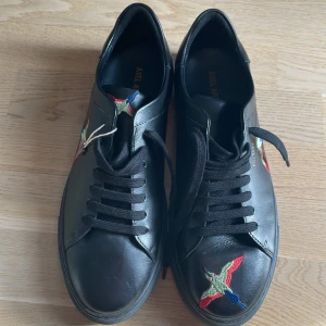 Svarta sneakers med broderad fågel - Snygga svarta sneakers från Axel Arigato med färgglada broderade fåglar på ovansidan. Skorna har klassisk snörning och en rund tå. Perfekta för en stilren look med en touch av färg.