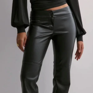 Svarta skinnbyxor  - Snygga svarta skinnbyxor med låg midja. Byxorna har en stilren design med knappar i midjan och en tight passform som framhäver figuren. Perfekta för en trendig look.