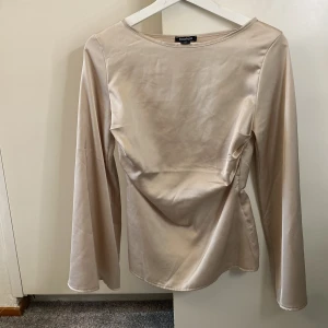 Beige långärmad topp från Boohoo - Säljer en elegant beige långärmad topp från Boohoo. Toppen har en fin drapering på framsidan och är tillverkad i ett glansigt material som ger en lyxig känsla. Perfekt för att klä upp en outfit! 💫super snygga breda ärmar🥰