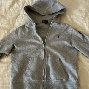 Grå hoodie från Polo Ralph Lauren - Säljer en stilren grå hoodie från Polo Ralph Lauren med dragkedja och broderad logga på bröstet. Tröjan har långa ärmar och en praktisk huva. Perfekt för en avslappnad look.
