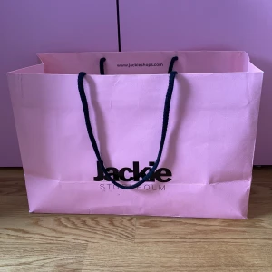 Jackie påse - Snygg rosa shoppingväska från Jackie Stockholm med svarta handtag. Perfekt för en dag på stan eller som en stilren accessoar. Väskan har en enkel och modern design.
