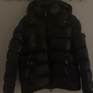 Svart dunjacka från Moncler - Säljer en stilren svart dunjacka från Moncler. Jackan har en glansig finish och är utrustad med dragkedja och flera praktiska fickor. Perfekt för kyliga dagar med sin varma och bekväma design.