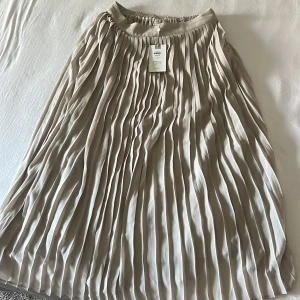Beige lång kjol från Vila - Säljer en elegant beige plisserad kjol från Vila, perfekt inför sommaren. Kjolen har en mjuk och flödande design med elastisk midja. Storlek 38, aldrig använd. Kan diskutera pris. Köpt för 400.
