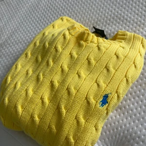 Gul stickad tröja från Ralph Lauren - Säljer en snygg gul stickad tröja från Ralph Lauren med kabelstickat mönster. Tröjan har långa ärmar och en broderad blå logotyp på bröstet. Perfekt för att lysa upp garderoben! Slim Fit