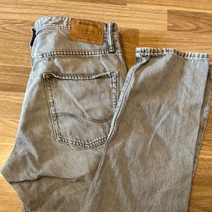 Gråa jeans Jack & Jones - Snygga grå jeans från Jack & Jones med klassisk femficksdesign och knappgylf. Perfekta för en avslappnad stil. Märkeslapp i läder baktill ger en extra touch. De har storlek W32 L34