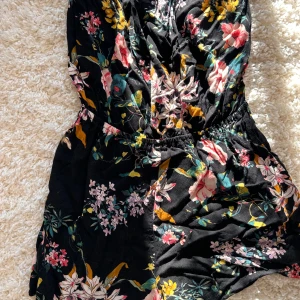 Svart blommig jumpsuit från Divided - Säljer en svart kort jumpsuit från Divided med ett färgglatt blommönster. Den är tillverkad i ett lätt och bekvämt material som ger en skön passform. Stl 38
