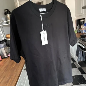 Svart t-shirt från Lanvin - Säljer en stilren svart t-shirt från Lanvin med broderad logga på bröstet. T-shirten är tillverkad i mjuk bomull och har en klassisk passform med korta ärmar. Perfekt för en minimalistisk look. Köpt för ca 3500kr men var i fel storlek.