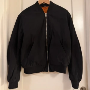 Svart bomberjacka med dragkedja - Snygg svart bomberjacka med dragkedja och ribbade muddar. Jackan har en klassisk design med en orange insida