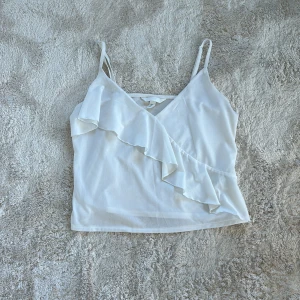 Vit linnetopp med volang - Säljer en söt vit linnetopp med smala axelband och volangdetalj framtill. Perfekt för varma sommardagar eller en avslappnad kvällslook. Toppen har en luftig och lätt passform.