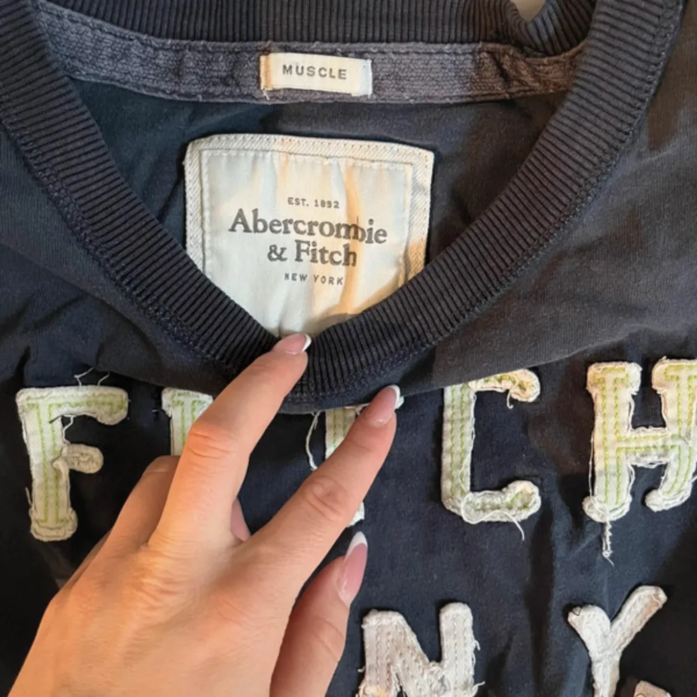 Säljer en mörkblå t-shirt från Abercrombie & Fitch med texten 'FITCH 8TH N.Y. ATHL' i slitna bokstäver på framsidan. . T-paidat.
