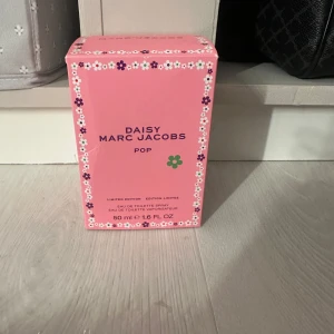 Daisy Marc Jacobs Pop Eau de Toilette - Limited edition Daisy Marc Jacobs Pop Eau de Toilette i en rosa flaska med dekorativa blommor på locket. Flaskan rymmer 50 ml och har en lekfull design med vita och lila blommor. Perfekt för den som älskar en fräsch och blommig doft.