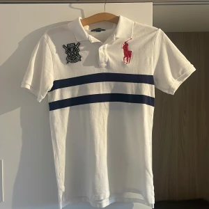 Polo Ralph Lauren piké - Vit pikétröja från Polo Ralph Lauren med två mörkblå horisontella ränder över bröstet. Stor röd logotyp och broderad klubbpatch framtill. Klassisk krage och korta ärmar. Perfekt för en sportig och stilren look.