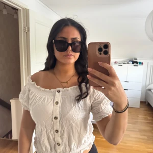 Vit offshoulder blus med knappar - Säljer en söt vit offshoulder blus med knappar framtill och korta puffärmar. Blusen har en croppad passform och resår i midjan för en bekväm känsla. Perfekt för varma dagar!