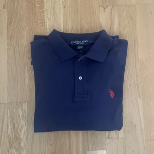 Marinblå pikétröja från U.S. Polo Assn. - Klassisk marinblå pikétröja från U.S. Polo Assn. med röd broderad logga på bröstet. Tröjan har krage, knappar framtill och är tillverkad i mjuk bomull. Perfekt för en clean och sportig stil.
