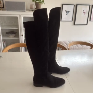 Svarta höga stövlar i mocka och textil - Snygga svarta overknee-stövlar med platt sula och rund tå. Gjorda i en mix av mocka och textil, med dragkedja på insidan för enkel påtagning. Perfekta för att lyfta din outfit under höst och vinter.