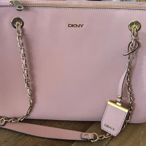 Ljusrosa axelväska från DKNY - Säljer en snygg ljusrosa axelväska från DKNY med guldfärgade kedjedetaljer och dragkedja upptill. Väskan har ett klassiskt och stilrent utseende med DKNY-logga både på utsidan och insidan. Perfekt för dig som vill ha en elegant accessoar. Helt oanvänd. Mått: 34*12*24cm Kommer inte till användning, därav säljs den. Kvitto finns ej kvar.