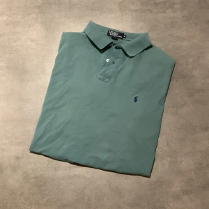 Grön pikétröja från Polo Ralph Lauren - Klassisk grön pikétröja från Polo Ralph Lauren med korta ärmar och krage. Tröjan har knappar vid halsen och den ikoniska broderade loggan i blått på bröstet. Perfekt för en avslappnad och stilren look. Tröjan är i storlek XL i herr. Tröjan är blekt som ni ser på prickarna. Bara att höra av sig vid funderingar eller frågor! Priset kan diskuteras!😀😀