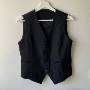 Svart kostymväst med knappar - Klassisk svart väst med tre knappar framtill och två fickor. Västen har en v-ringad design och är figursydd för en stilren look. 