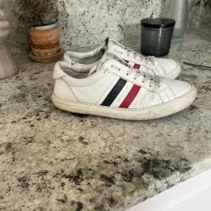 Vita sneakers från Moncler  - Säljer ett par vita Moncler sneakers med klassiska ränder i rött och blått på sidan. Skorna har snörning, rund tå och platt sula. Perfekta för dig som gillar stilrena och sportiga skor med en touch av lyx. Skick 6/10 pris kan diskuteras!