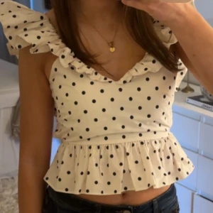 Vit prickig volangtopp - Söt vit topp med svarta prickar och volangdetaljer vid axlar och nederkant. Toppen har en figurnära passform upptill och är ärmlös med breda volangärmar. Perfekt för en somrig look