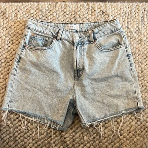 Ljusblå jeansshorts med fransar - Säljer ett par ljusblå jeansshorts som är lite längre med råa fransade benslut och klassisk femficksdesign. Shortsen har hög midja och en avslappnad passform, perfekta för sommardagar.