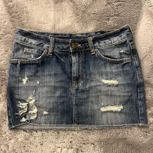 Mörkblå jeanskjol med slitningar - Säljer en snygg mörkblå jeanskjol med slitna detaljer och rå kant nertill. Kjolen har klassiska fickor fram och bak samt bälteshällor och knappstängning. Perfekt för en cool och avslappnad stil.