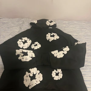 Denim Tears Hoodie - Säljer en svart hoodie med stora vita blommor tryckta över hela tröjan. Hoodien har en klassisk passform med huva och långa ärmar. Perfekt för dig som vill sticka ut lite extra med ett unikt mönster.