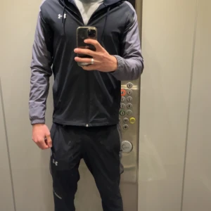 Svart tränings dress från Under Armour - Säljer en svart tränings dress från Under Armour med dragkedja och huva. Tröjan har långa ärmar i en mörkgrå nyans och är tillverkad i ett syntetmaterial som passar perfekt för träning. Reflekterande detaljer på ärmar och ben. reflektive och finns drag kedja på benet där du kan lösa upp Storlek S jacka M byxor