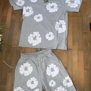 Denim tears tracksuit  - SELGER FRA NORGE ER 900 NORSKE KRONER! Snygg grå t-shirt från Denim Tears med stora vita blommiga tryck över hela tröjan. T-shirten har rund hals, korta ärmar och en relaxed fit. Perfekt för dig som gillar streetwear och vill sticka ut med ett unikt mönster. BÅDE SHORTS OG T-SKJORTE