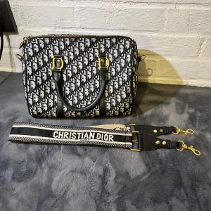 Christian Dior svartvit handväska - Snygg handväska från Christian Dior i svartvitt mönster med tydlig logga. Väskan har guldfärgade metalldetaljer, svarta handtag i skinn och en bred axelrem med Christian Dior-tryck. Tillverkad i bomullstyg med skinninslag och dragkedja upptill.