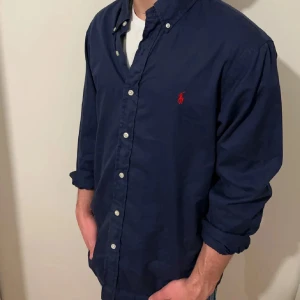 Mörkblå skjorta från Ralph Lauren - Snygg mörkblå skjorta från Ralph Lauren i slim fit-modell. Skjortan har klassisk krage, knäppning framtill och det ikoniska röda broderade logot på bröstet. Tillverkad i bomullstwill som känns både mjuk och stadig. Perfekt för dig som gillar stilrena och tidlösa plagg.