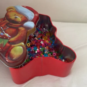 Färgglada pärlor i julburk - En stor samling färgglada plastpärlor i olika former och färger, perfekt för att göra egna armband, halsband eller andra smycken. Pärlorna levereras i en röd plåtburk med julmotiv och nallebjörn på locket.