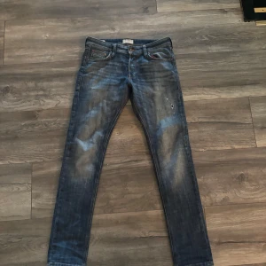 Slim Glenn blå jeans med slitningar - Snygga blå jeans från Slim Glenn med slitna detaljer och tvättad look. Modellen är slim fit med klassiska fem fickor och orangea sömmar. Jeansen har en smal passform och är perfekta för dig som gillar en modern stil. Storlek 30 midja och 34 längd.