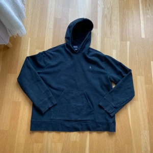 Svart hoodie från Polo Ralph Lauren - Mörkblå hoodie från Polo Ralph Lauren med klassisk huva och stor magficka. Tröjan har diskret logga på bröstet och är tillverkad i mjuk bomullsblandning. Perfekt för en chill och stilren look. Den är i bra skick förutom en liten fläck på ena armslutet