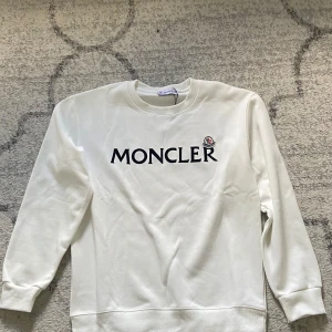 Vit sweatshirt från Moncler - Säljer en stilren vit sweatshirt från Moncler med svart logotyptryck på bröstet och broderad Moncler-patch. Tröjan har rund halsringning och långa ärmar. Perfekt för dig som gillar klassisk och enkel design.
