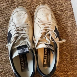 Vita sneakers från Golden Goose Ballstar - Säljer ett par vita Golden Goose Ballstar sneakers med svarta detaljer och stjärna på sidan. Skorna har snörning, rund tå och platt sula. Klassisk vintage-look med slitna detaljer och svart hälparti. Perfekta för dig som gillar streetstyle.