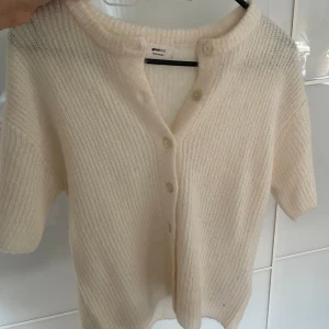 Vit kofta från Gina Tricot - Säljer en mjuk och ribbstickad vit kofta från Gina Tricot i ullblandning. Koftan har korta ärmar, v-ringning och knäppning framtill med fyra knappar. Perfekt för lager-på-lager och enkel att matcha.