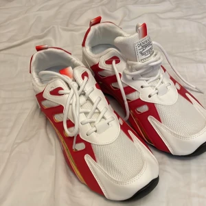 Vita och röda sneakers med gulddetaljer - Snygga sneakers i vitt och rött med coola gulddetaljer på sidorna. Ovandelen är i mesh och syntet med vita snören och svart sula. Skorna har en sportig vibe och sticker ut med sin färgkombination och moderna design.