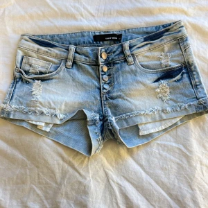Lågmidjade jeansshorts från Tally Weijl - Säljer ett par ljusblå lågmidjade jeansshorts från Tally Weijl med slitna detaljer och fransig kant. Shortsen har fyra knappar framtill, klassiska fickor och en cool, avslappnad look. Perfekta för varma dagar och enkel att matcha med olika toppar.