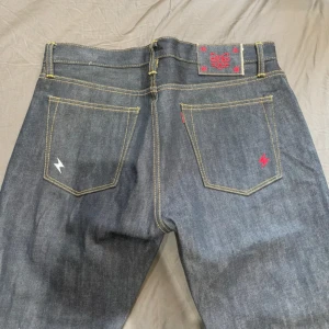 Mbbcar vintage Mörkblå japanska jeans med blixt-detalj - Riktigt snygga mörkblå jeans med raka ben och kontrastsömmar. Bakfickorna har broderade blixtar i vitt och rött, och det finns en röd patch med japanska tecken i midjan. Jeansen är tillverkade i kraftigt denimtyg.