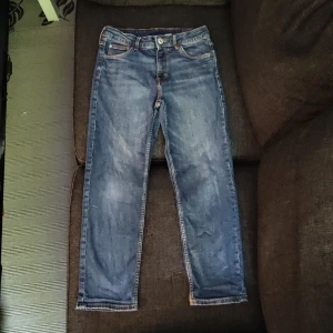 Blå jeans med relaxed fit från H&M - Säljer ett par blå jeans från H&M med relaxed fit och justerbar midja. Byxorna har klassisk femficksdesign, avsmalnande ben och synliga kontrastsömmar. Perfekta för en avslappnad stil. Dock saknas några knappar på back fickorna men annars är det inga större slitage.